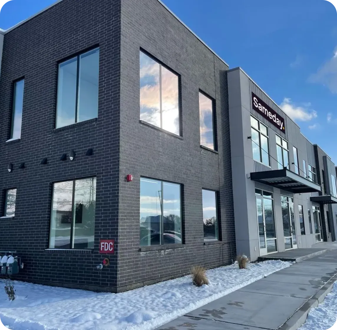 Modern two-story commercial building with dark brick and large windows, snow on ground, and a sign reading 'Sameday.'