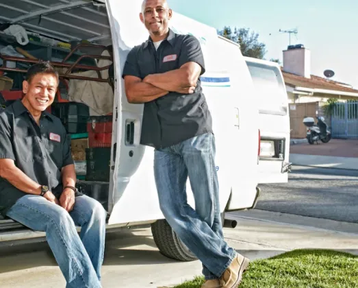 Two men wearing black work shirts and jeans standing and sitting by a white van parked in a driveway.